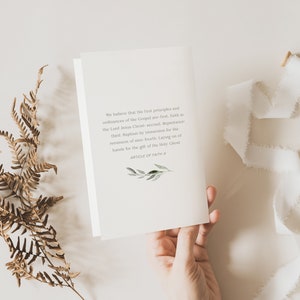 May include: A white card with text from the Article of Faith #4, held by a hand. The text is in a serif font and includes a small green and white botanical illustration. A white ribbon and dried fern are also in the image.