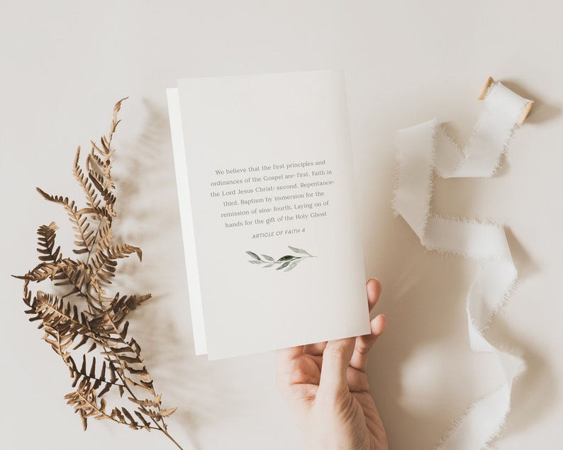 May include: A white card with text from the Article of Faith #4, held by a hand. The text is in a serif font and includes a small green and white botanical illustration. A white ribbon and dried fern are also in the image.