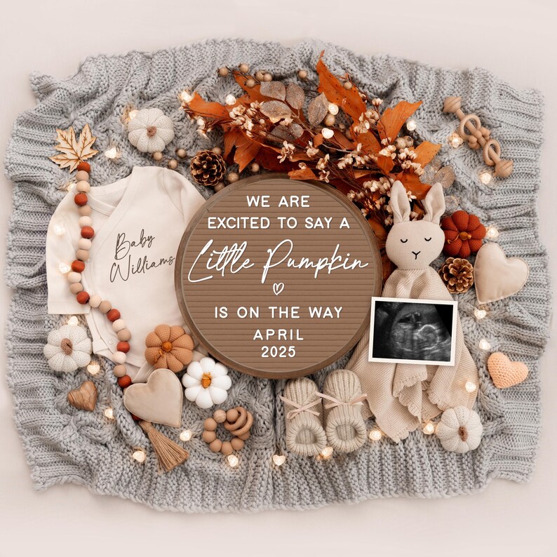 May include: A wooden sign with the text "We are excited to say a little pumpkin is on the way April 2025" surrounded by autumn-themed baby items, including a onesie, booties, a pumpkin, and a stuffed bunny.