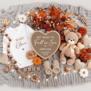 Puede incluir: Un letrero de madera en forma de coraz&oacute;n con el texto "Get ready to Fall in Love" y "Arriving August 2025". El letrero est&aacute; rodeado de decoraciones de oto&ntilde;o, incluyendo un body de beb&eacute; con el texto "Baby Oliver", un osito de peluche y un par de botines.