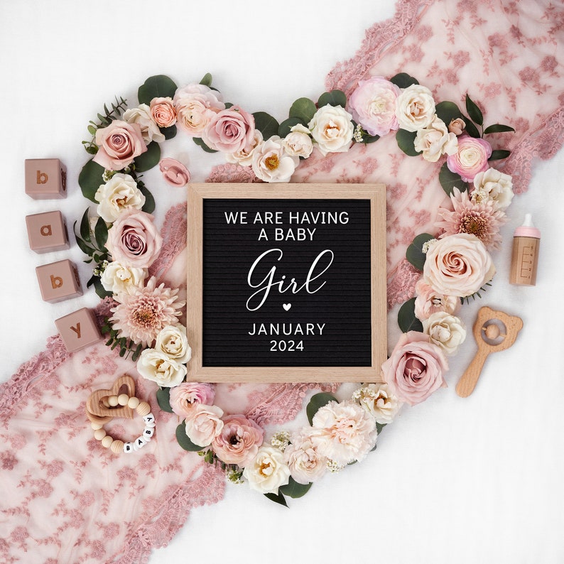May include: A pink and white floral arrangement in the shape of a heart with a letter board that reads "We are having a baby girl January 2024". There are also baby items such as a rattle, a bottle, and blocks that spell out "baby".