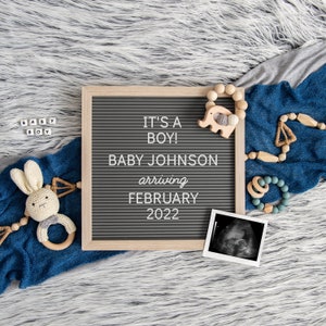 May include: A gray letter board with the text "It's a Boy! Baby Johnson arriving February 2022".  The board is surrounded by a blue blanket, a wooden elephant toy, and a wooden baby rattle.