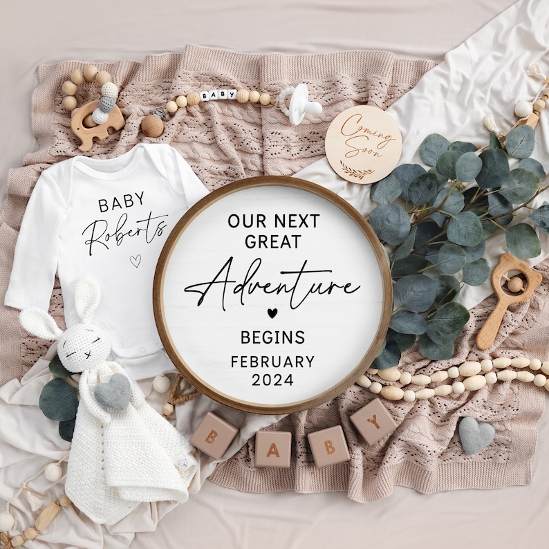 May include: A white baby onesie with the text "BABY Roberts" and a heart. A wooden round sign with the text "OUR NEXT GREAT Adventure BEGINS FEBRUARY 2024" and a heart. A wooden sign with the text "Coming Soon". A wooden toy elephant, a wooden rattle, and a wooden bead necklace with the word "BABY" on it.
