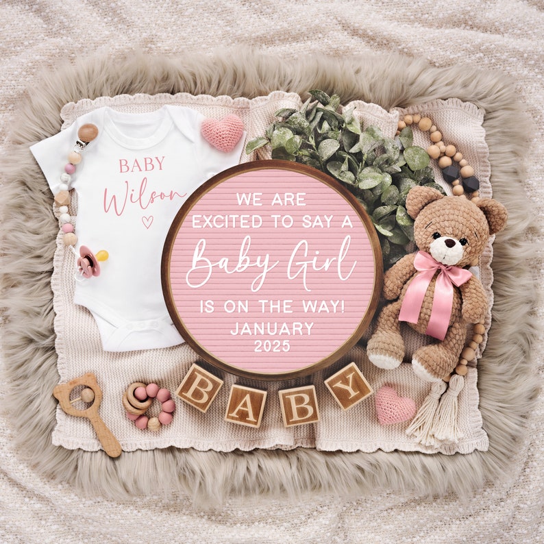 K&ouml;nnte beinhalten: Rosa-wei&szlig;e Baby-Ank&uuml;ndigung mit einem Holzschild, auf dem steht "We are excited to say a Baby Girl is on the way! January 2025". Es gibt einen wei&szlig;en Strampler mit dem Text "Baby Wilson" und einen Teddyb&auml;r mit einer rosa Schleife.
