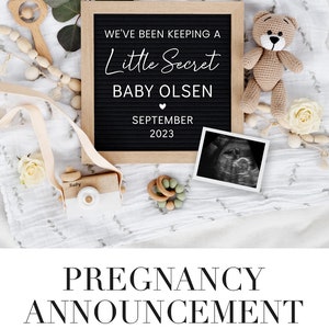 K&ouml;nnte beinhalten: Ein schwarzes Buchstabenbrett mit wei&szlig;em Text, der eine Schwangerschaft ank&uuml;ndigt. Der Text lautet "We've been keeping a little secret, Baby Olsen, September 2023". Das Brett ist von babybezogenen Gegenst&auml;nden umgeben, darunter ein Teddyb&auml;r, eine Spielzeugkamera aus Holz und ein Ultraschallbild.