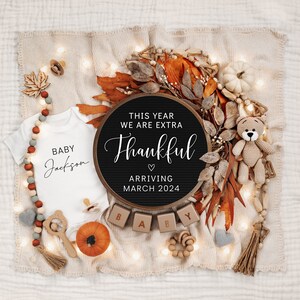 May include: A black letter board with the text "This year we are extra thankful" and a heart symbol. Below the text is "Arriving March 2024". The letter board is surrounded by fall-themed decorations, including a white onesie with the text "Baby Jackson", a small pumpkin, and wooden blocks that spell out "Baby".