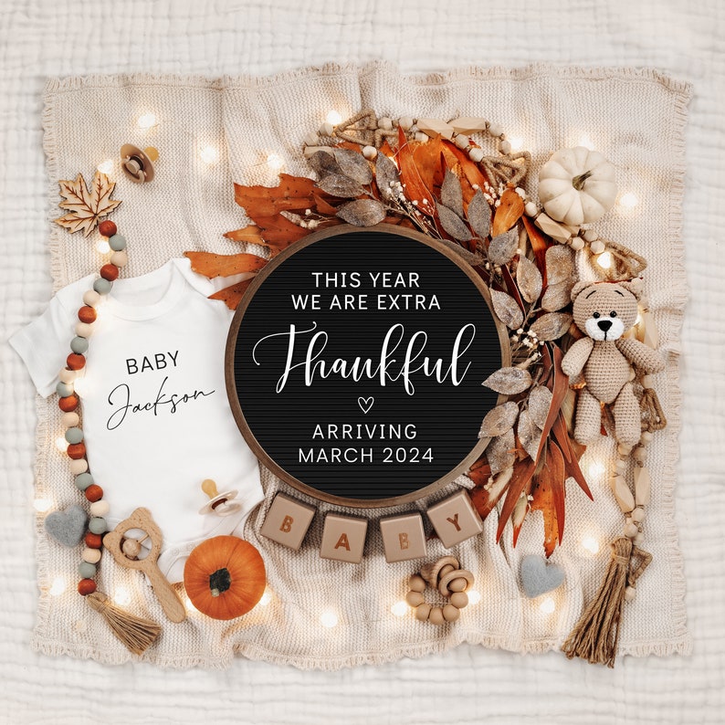 May include: A black letter board with the text "This year we are extra thankful" and a heart symbol. Below the text is "Arriving March 2024". The letter board is surrounded by fall-themed decorations, including a white onesie with the text "Baby Jackson", a small pumpkin, and wooden blocks that spell out "Baby".
