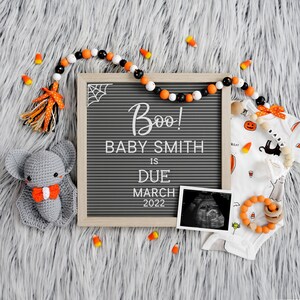 May include: A gray letter board with the text "Boo! Baby Smith is due March 2022".  A gray crocheted bat, an orange and white beaded garland, and a white onesie with a Halloween theme are on a gray fuzzy surface.