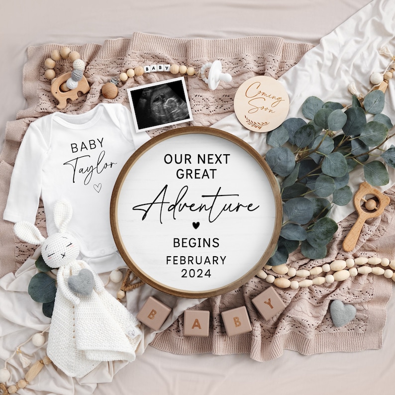 May include: A wooden sign with the text "Our Next Great Adventure Begins February 2024" surrounded by baby items, including a onesie that says "Baby Taylor", a wooden elephant, a wooden rattle, and a wooden block that spells "BABY".
