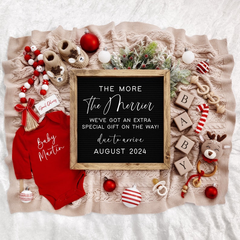 Puede incluir: Un mameluco rojo para beb&eacute; con el texto "Baby Martin" en &eacute;l, rodeado de decoraciones navide&ntilde;as, incluyendo un letrero de pizarra que dice "The More The Merrier We've Got An Extra Special Gift On The Way! Due To Arrive August 2024".