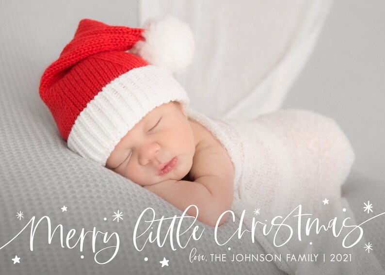 May include: A newborn baby wearing a red and white Santa hat sleeps on a gray blanket. The text "Merry little Christmas" is written in white cursive script with stars. The text "love, THE JOHNSON FAMILY | 2021" is written below in white.