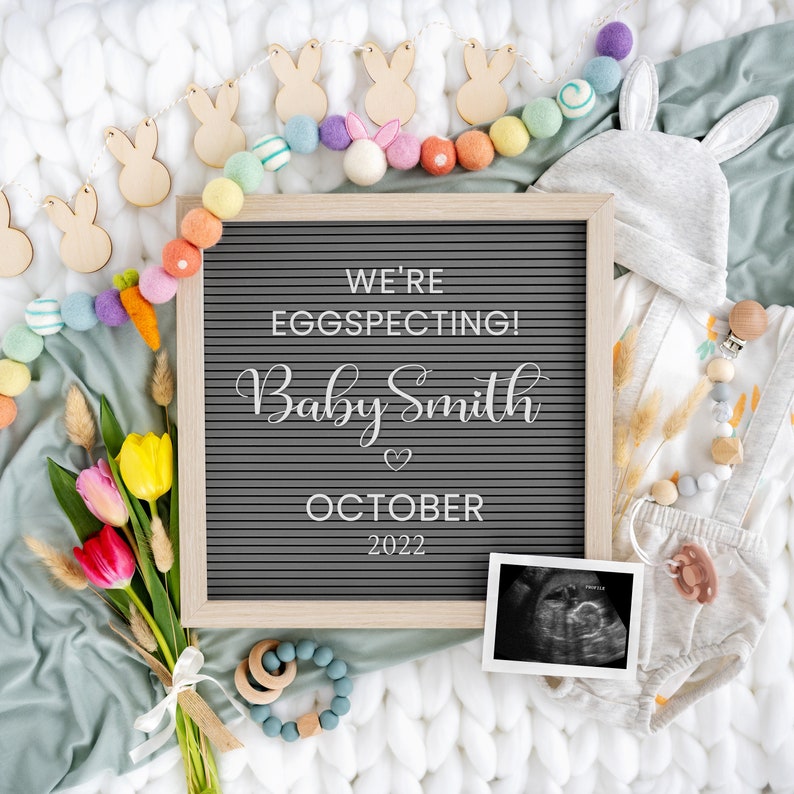 May include: A letter board with the text "We're Eggspecting! Baby Smith October 2022" surrounded by a garland of colourful felt balls and wooden bunny shapes. A baby onesie, a dummy, and an ultrasound image are also included in the image.