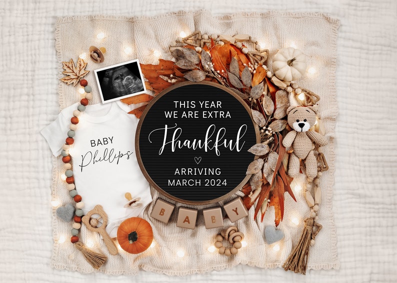 May include: A fall-themed baby announcement with a black chalkboard sign that reads "This year we are extra thankful arriving March 2024". The sign is surrounded by fall leaves, a small stuffed bear, and a baby onesie that reads "Baby Phillips".