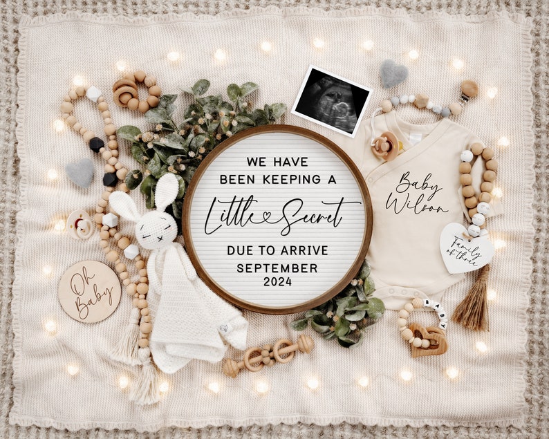 May include: A wooden sign with the text "We have been keeping a little secret due to arrive September 2024" surrounded by a white blanket, a baby onesie with the text "Baby Wilson", a knitted bunny toy, and wooden beads.