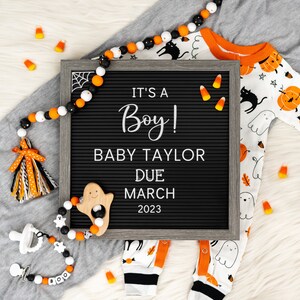 May include: A black letter board with the text "It's a Boy! Baby Taylor Due March 2023".  A white and orange baby onesie with a ghost and pumpkin design is on a white surface with orange and white candy corn and a black and white beaded garland.