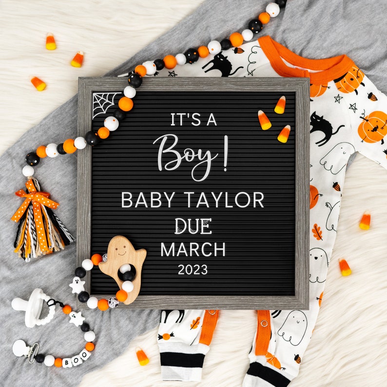 May include: A black letter board with the text "It's a Boy! Baby Taylor Due March 2023".  A white and orange baby onesie with a ghost and pumpkin design is on a white surface with orange and white candy corn and a black and white beaded garland.