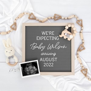 May include: A gray letter board with the text "We're expecting Baby Wilson arriving August 2022". The board is surrounded by a white fabric and wooden beads with a small wooden elephant toy.
