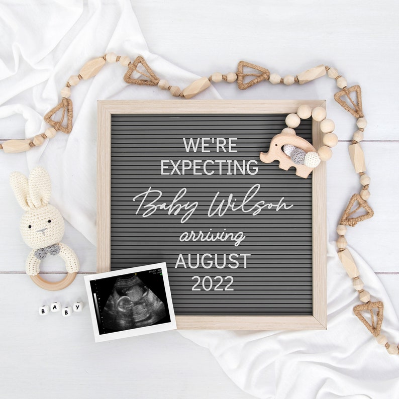 May include: A gray letter board with the text "We're expecting Baby Wilson arriving August 2022". The board is surrounded by a white fabric and wooden beads with a small wooden elephant toy.