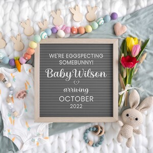 May include: A gray letter board with the text "We're eggspecting somebunny! Baby Wilson arriving October 2022". The board is surrounded by pastel colored felt balls, wooden bunny cutouts, and a bouquet of yellow and red tulips.