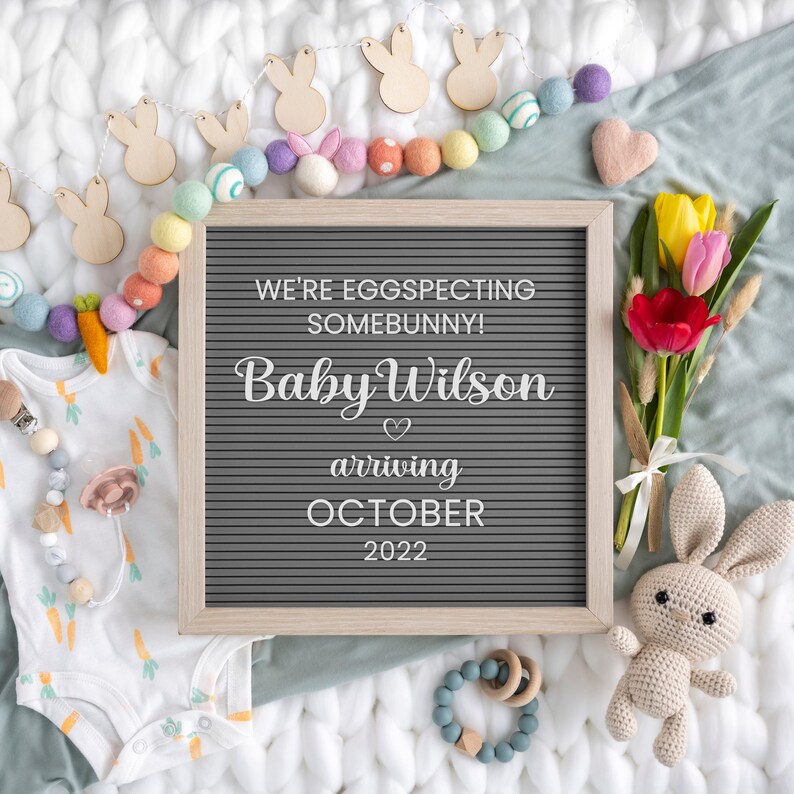 May include: A gray letter board with the text "We're eggspecting somebunny! Baby Wilson arriving October 2022". The board is surrounded by pastel colored felt balls, wooden bunny cutouts, and a bouquet of yellow and red tulips.