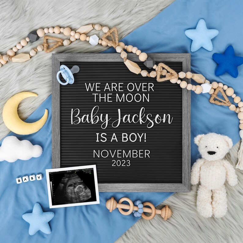 May include: A black letter board with the text "We are over the moon Baby Jackson is a boy! November 2023". The board is surrounded by a blue blanket, a yellow crescent moon, a white cloud, a pacifier, a teddy bear, a wooden rattle, and a sonogram image.