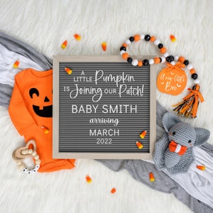 May include: A gray letter board with the text "A little Pumpkin is Joining our Patch! Baby Smith arriving March 2022".  There is a small orange pumpkin with a black face and a gray bat with a bow tie.  The background is white with orange and white candy corn scattered around.