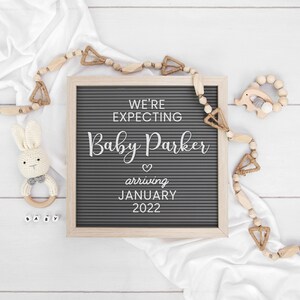 May include: A gray letter board with the text "We're expecting Baby Parker arriving January 2022". The board is surrounded by wooden beads and a white fabric.