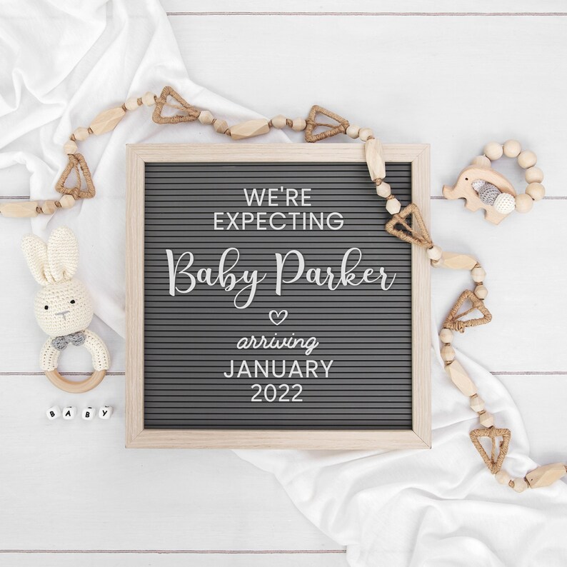 May include: A gray letter board with the text "We're expecting Baby Parker arriving January 2022". The board is surrounded by wooden beads and a white fabric.