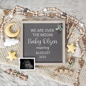 Peut inclure: Un tableau d'affichage en lettres grises avec le texte "We are over the moon! Baby Olsen arriving August 2023". Le tableau est entour&eacute; d'une vari&eacute;t&eacute; de d&eacute;corations sur le th&egrave;me des b&eacute;b&eacute;s, notamment une &eacute;toile jaune, un nuage blanc, une lune croissante jaune, une sucette, des perles en bois et un hochet en bois.