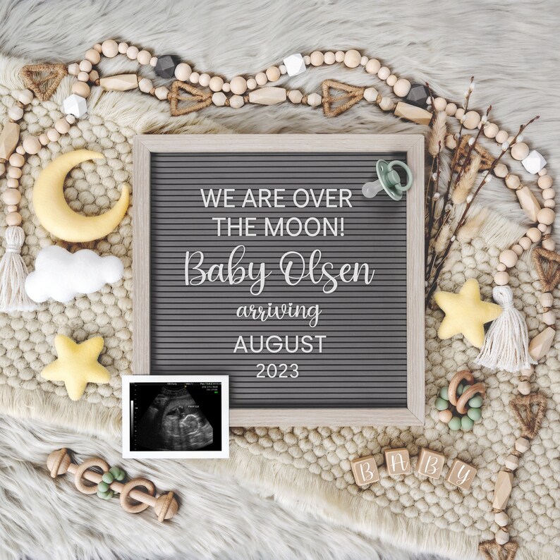 Peut inclure: Un tableau d'affichage en lettres grises avec le texte "We are over the moon! Baby Olsen arriving August 2023". Le tableau est entour&eacute; d'une vari&eacute;t&eacute; de d&eacute;corations sur le th&egrave;me des b&eacute;b&eacute;s, notamment une &eacute;toile jaune, un nuage blanc, une lune croissante jaune, une sucette, des perles en bois et un hochet en bois.