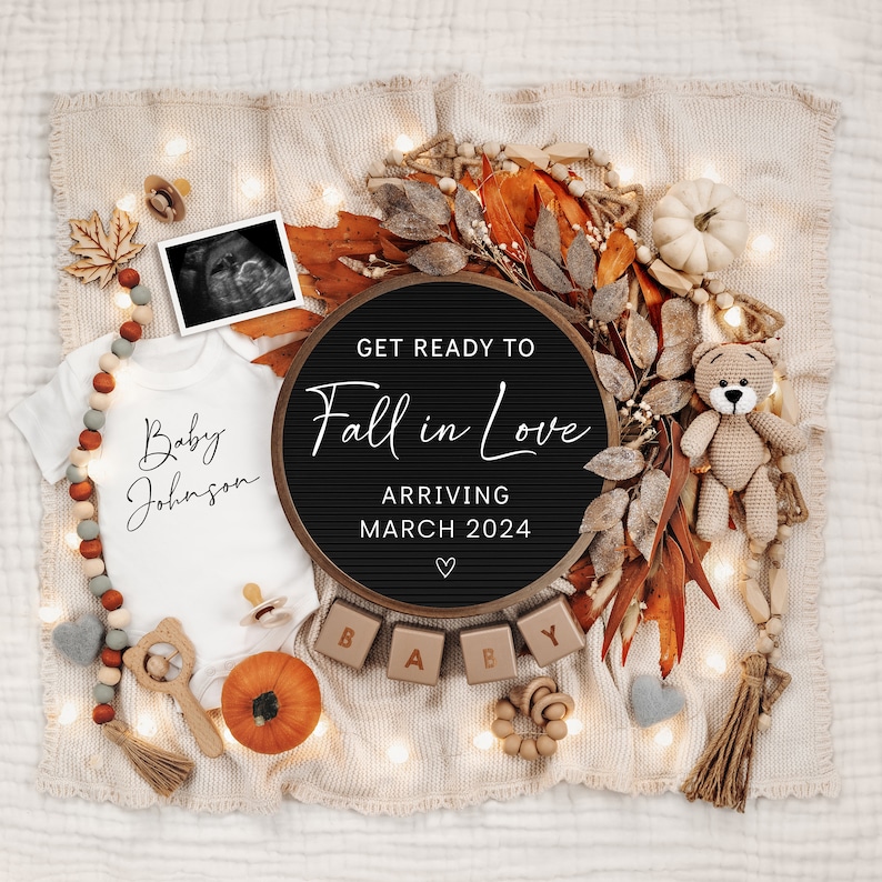 May include: A black letter board with the text "Get Ready to Fall in Love" and "Arriving March 2024" surrounded by autumn-themed decorations, including a white onesie with the text "Baby Johnson", a pumpkin, and a crocheted teddy bear.