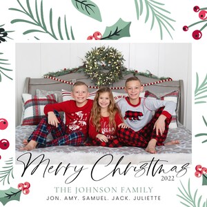 May include: A Christmas card featuring a family of five in matching pajamas. The family is sitting on a bed with a Christmas wreath behind them. The card has a green and red holly design with the text "Merry Christmas 2022" and "THE JOHNSON FAMILY JON. AMY. SAMUEL. JACK. JULIETTE".