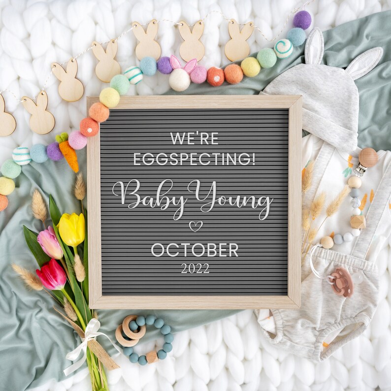 May include: A letter board with the text "We're Eggspecting! Baby Young October 2022" surrounded by a garland of colourful felt balls and wooden bunny shapes. A baby outfit, a hat, and a wooden toy are also in the image.
