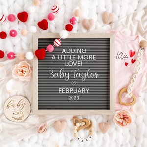 May include: A letter board with the text "Adding a little more love! Baby Taylor February 2023" surrounded by pink and white felt balls, a pink onesie with the word "love" and a wooden heart, and a wooden baby rattle.