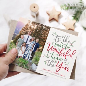 May include: A Christmas card with a photo of a family of six standing in a forest. The card has the text "It's the most Wonderful Time of the Year" in red and green lettering with green foliage accents. The text "The Smith Family 20XX" is printed below the main text with the names of the family members listed below.