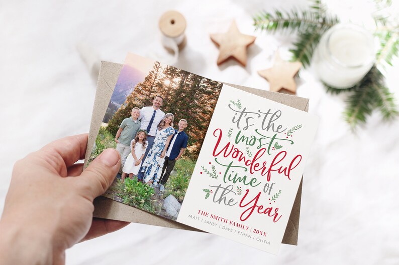 May include: A Christmas card with a photo of a family of six standing in a forest. The card has the text "It's the most Wonderful Time of the Year" in red and green lettering with green foliage accents. The text "The Smith Family 20XX" is printed below the main text with the names of the family members listed below.