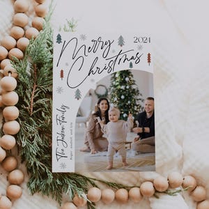Pu&ograve; includere: Un biglietto di Natale con sfondo bianco e un design festivo che presenta una famiglia di tre persone davanti a un albero di Natale decorato. Il testo "Merry Christmas 2021" &egrave; scritto in corsivo nero con accenti di fiocco di neve bianco e albero di pino verde. La famiglia &egrave; in piedi davanti a un albero di Natale decorato. Il biglietto include anche il testo "The Jackson Family Roger, anabell, and lyla".