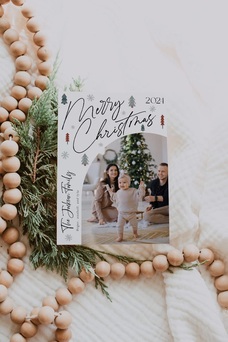 Pu&ograve; includere: Un biglietto di Natale con sfondo bianco e un design festivo che presenta una famiglia di tre persone davanti a un albero di Natale decorato. Il testo "Merry Christmas 2021" &egrave; scritto in corsivo nero con accenti di fiocco di neve bianco e albero di pino verde. La famiglia &egrave; in piedi davanti a un albero di Natale decorato. Il biglietto include anche il testo "The Jackson Family Roger, anabell, and lyla".