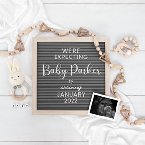 May include: A gray letter board with the text "We're expecting Baby Parker arriving January 2022". The board is surrounded by wooden beads and a white blanket. There is a small wooden elephant toy and a crocheted bunny toy with a rattle.