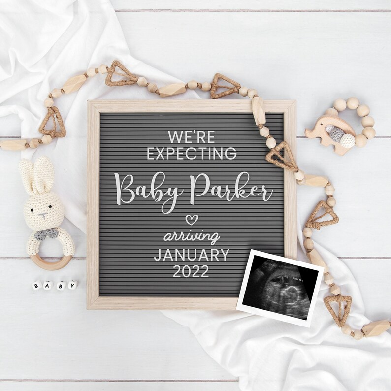 May include: A gray letter board with the text "We're expecting Baby Parker arriving January 2022". The board is surrounded by wooden beads and a white blanket. There is a small wooden elephant toy and a crocheted bunny toy with a rattle.