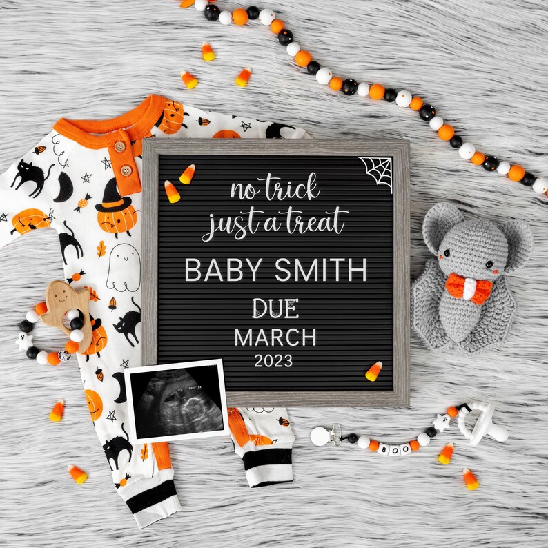 K&ouml;nnte beinhalten: Ein schwarzes Buchstabenbrett mit dem Text "no trick just a treat Baby Smith Due March 2023". Ein wei&szlig;-oranger Baby-Strampler mit einem schwarz-wei&szlig;en Halloween-Druck, eine graue geh&auml;kelte Fledermaus und ein schwarz-wei&szlig;er Schnuller mit dem Wort "BOO" darauf sind um das Buchstabenbrett herum angeordnet.