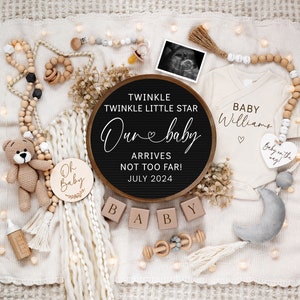 May include: A black letter board with the text "Twinkle Twinkle Little Star Our Baby Arrives Not Too Far! July 2024".  The letter board is surrounded by a variety of baby items, including a white onesie with the text "Baby Williams", a wooden rattle, a stuffed animal, and a wooden sign that says "Oh Baby".