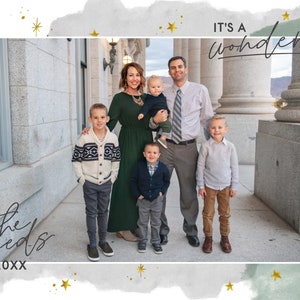 May include: A family of six poses in front of a building with columns. The family is standing on a sidewalk. The parents are holding a baby and a toddler. The two older children are standing on either side of the parents. The text "It's a wonderful life" is written in cursive at the top of the image. The text "Love, the Steeds 20XX" is written in cursive at the bottom of the image.