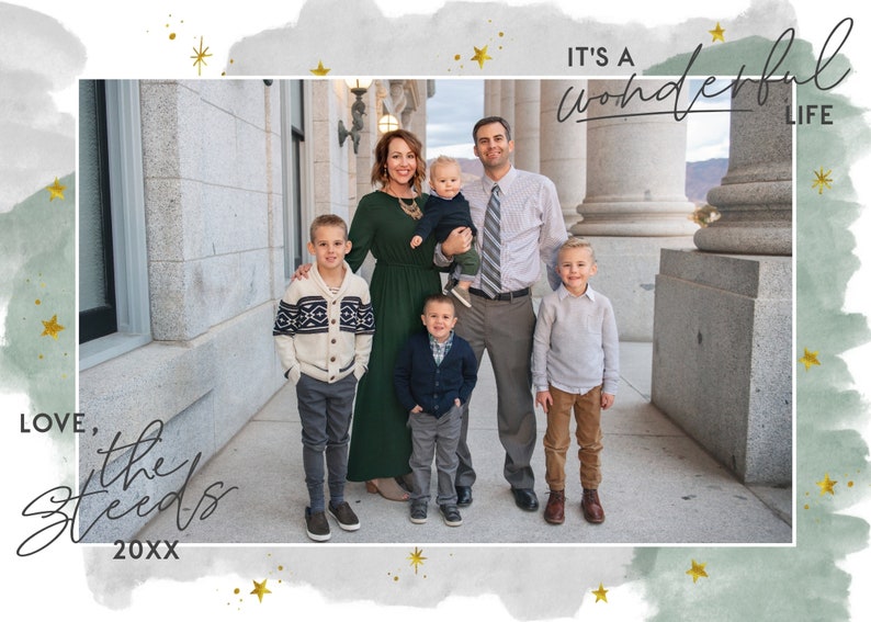 May include: A family of six poses in front of a building with columns. The family is standing on a sidewalk. The parents are holding a baby and a toddler. The two older children are standing on either side of the parents. The text "It's a wonderful life" is written in cursive at the top of the image. The text "Love, the Steeds 20XX" is written in cursive at the bottom of the image.