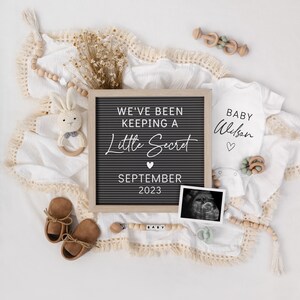 May include: A white blanket with a letter board that reads "We've Been Keeping A Little Secret September 2023". There is a white onesie with the text "Baby Wilson" and a black and white ultrasound image. The blanket is decorated with wooden beads, a wooden toy, and a stuffed bunny.