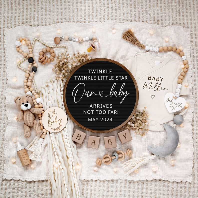 May include: A black letter board with the text "Twinkle Twinkle Little Star Our Baby Arrives Not Too Far! May 2024".  The letter board is surrounded by a white blanket, wooden blocks spelling "BABY", a wooden baby rattle, a crocheted teddy bear, a wooden sign with the text "Oh Baby", a white onesie with the text "Baby Miller", and a wooden heart with the text "Baby on the way".