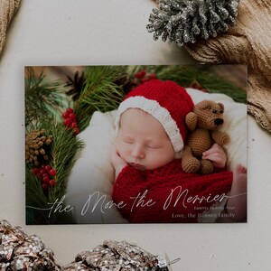 May include: A Christmas card featuring a sleeping baby wearing a red and white Santa hat and holding a brown teddy bear. The card has the text "The More the Merrier" and "Twenty Twenty-Four" in a cursive font. The text "Love, The Hansen Family" is printed below.