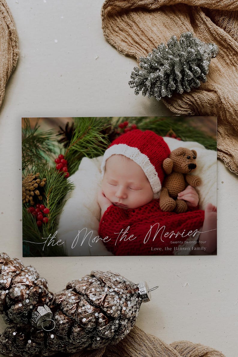 May include: A Christmas card featuring a sleeping baby wearing a red and white Santa hat and holding a brown teddy bear. The card has the text "The More the Merrier" and "Twenty Twenty-Four" in a cursive font. The text "Love, The Hansen Family" is printed below.