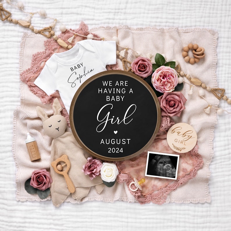 May include: A pink and white blanket with a black chalkboard sign that says "We are having a baby girl August 2024". There is a white onesie with the name "Sophia" on it, a wooden baby girl sign, a stuffed bunny, a pacifier, and a baby ultrasound picture.