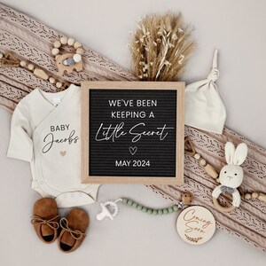 Puede incluir: Un anuncio de nacimiento con un tablero de letras que dice "We've been keeping a little secret May 2024". Hay un body blanco con el texto "Baby Jacobs" y un gorro de punto marr&oacute;n. El anuncio tambi&eacute;n incluye un letrero de madera que dice "Coming Soon" y un sonajero de madera con un conejito adjunto.